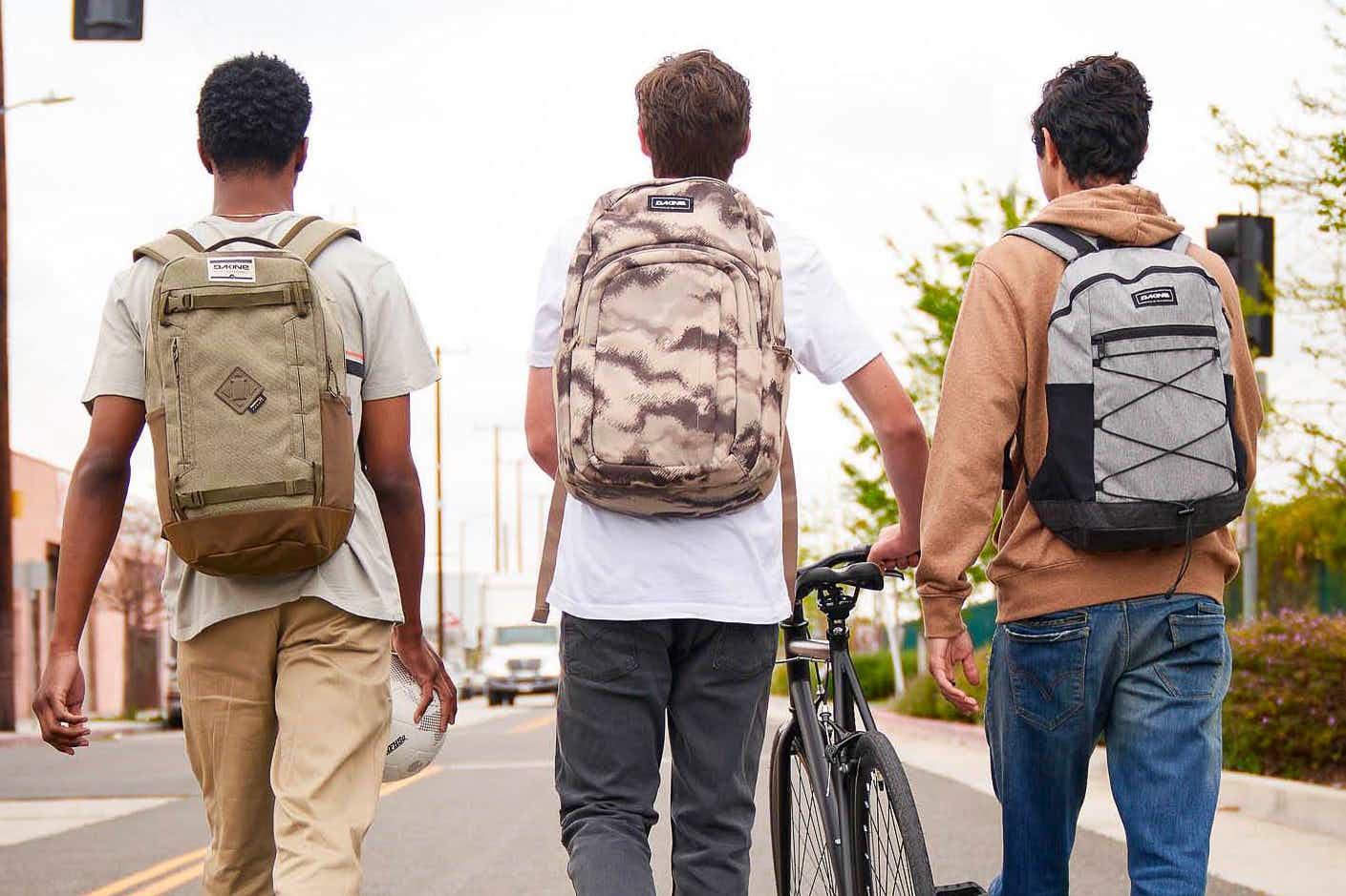 Three kids wearing Dakine backpacks walking together down a street, one of them pushing a bike.