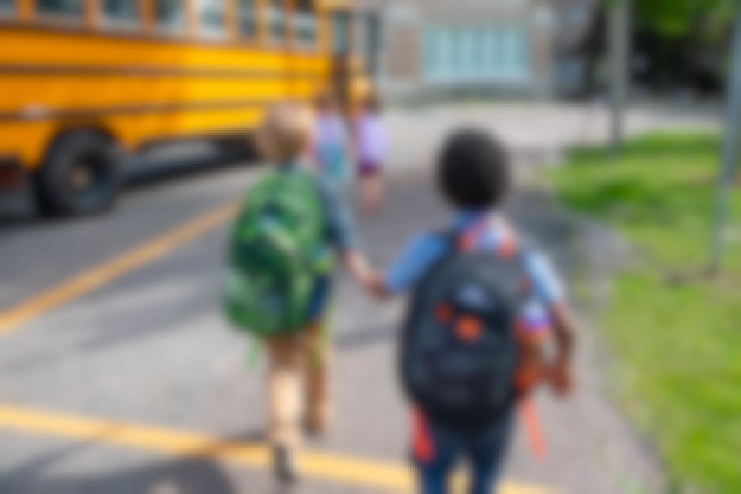 Two kids wearing High Sierra backpacks, holding hands and walking toward a school bus.
