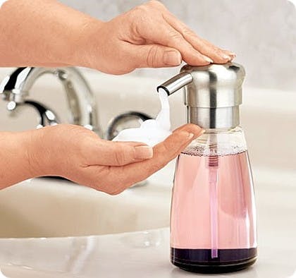 A person pushing down on a soap dispenser with one hand while it dispenses foaming soap into the person's hand.