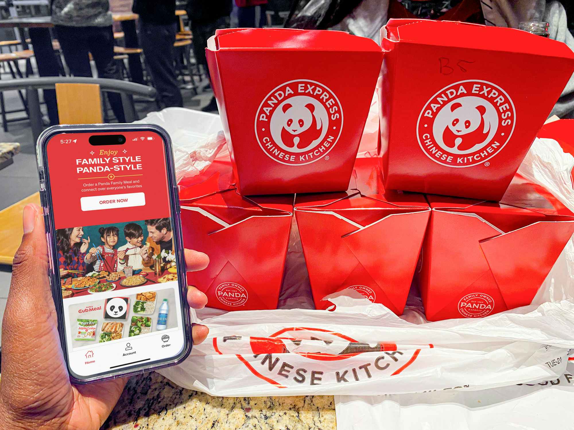 Someone holding a phone displaying the Panda Express app's main page featuring the family style meal next to some takeout boxes of food on a table at Panda Express