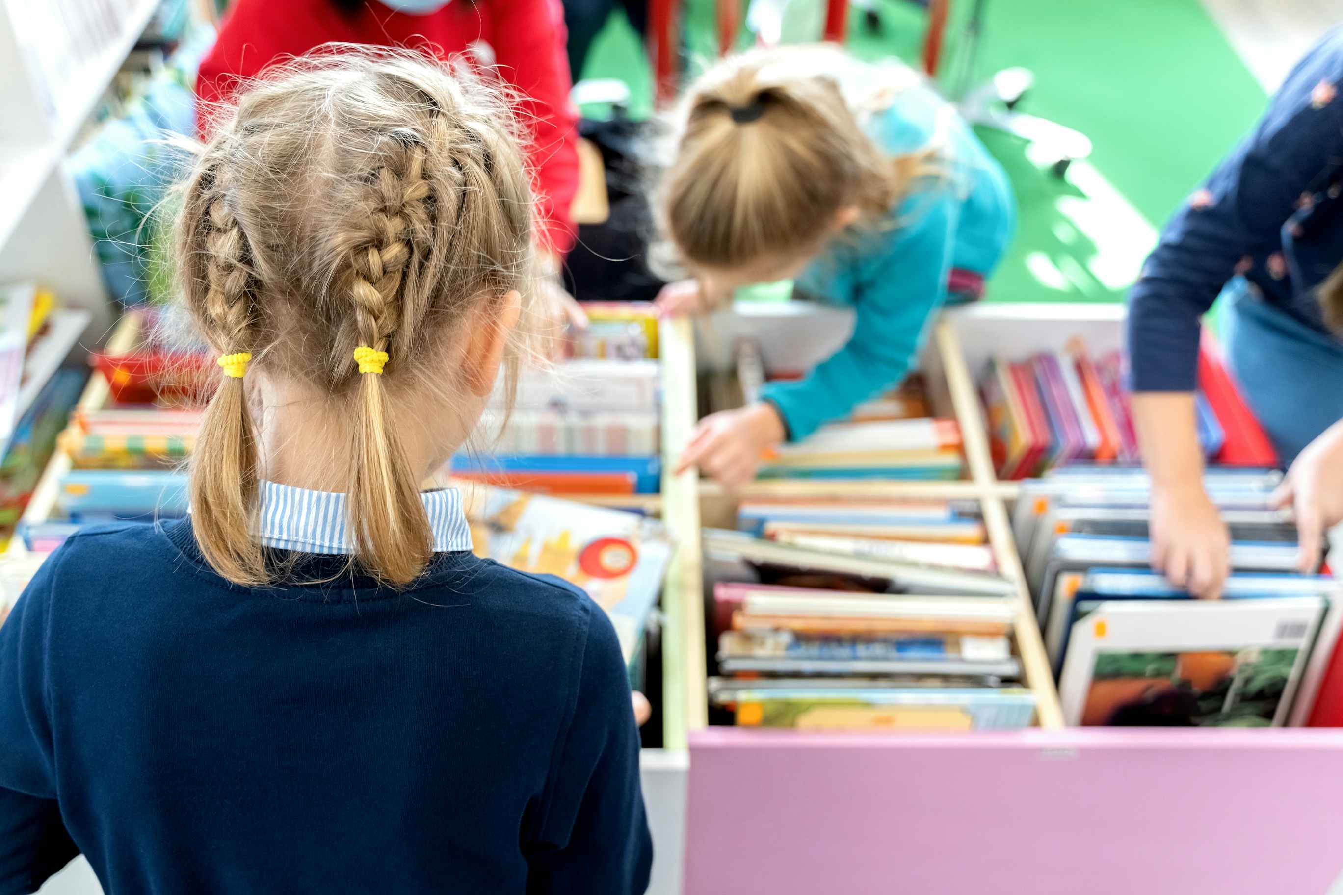 A child with French braids, facing away from the camera, looking at a bin full of books that other children are looking through.