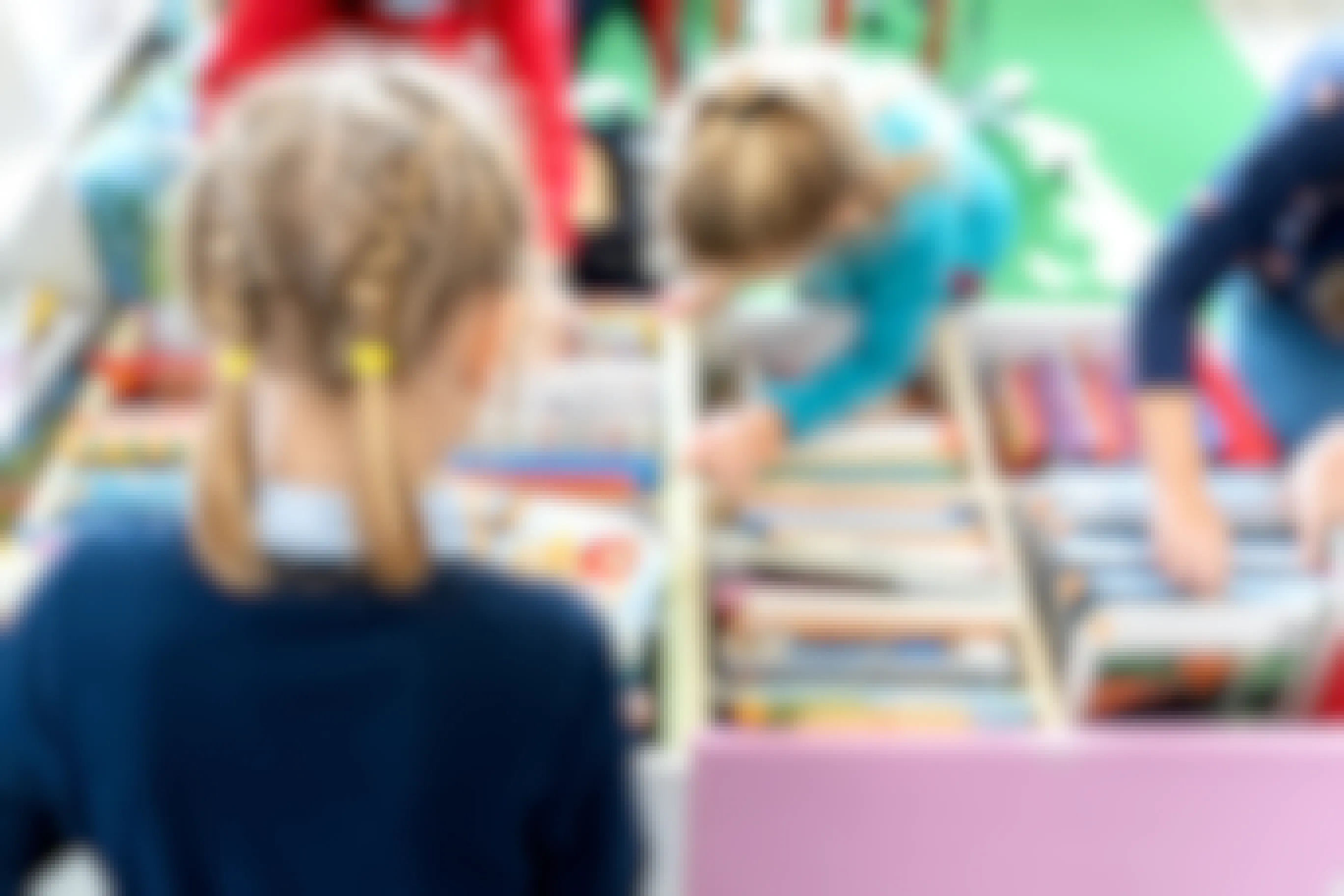 A child with French braids, facing away from the camera, looking at a bin full of books that other children are looking through.