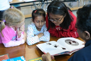 Three kids reading a children's book together at a desk.