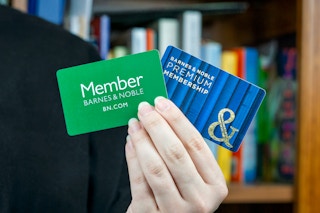someone standing near a bookshelf, holding up a free and premium barnes and noble membership card
