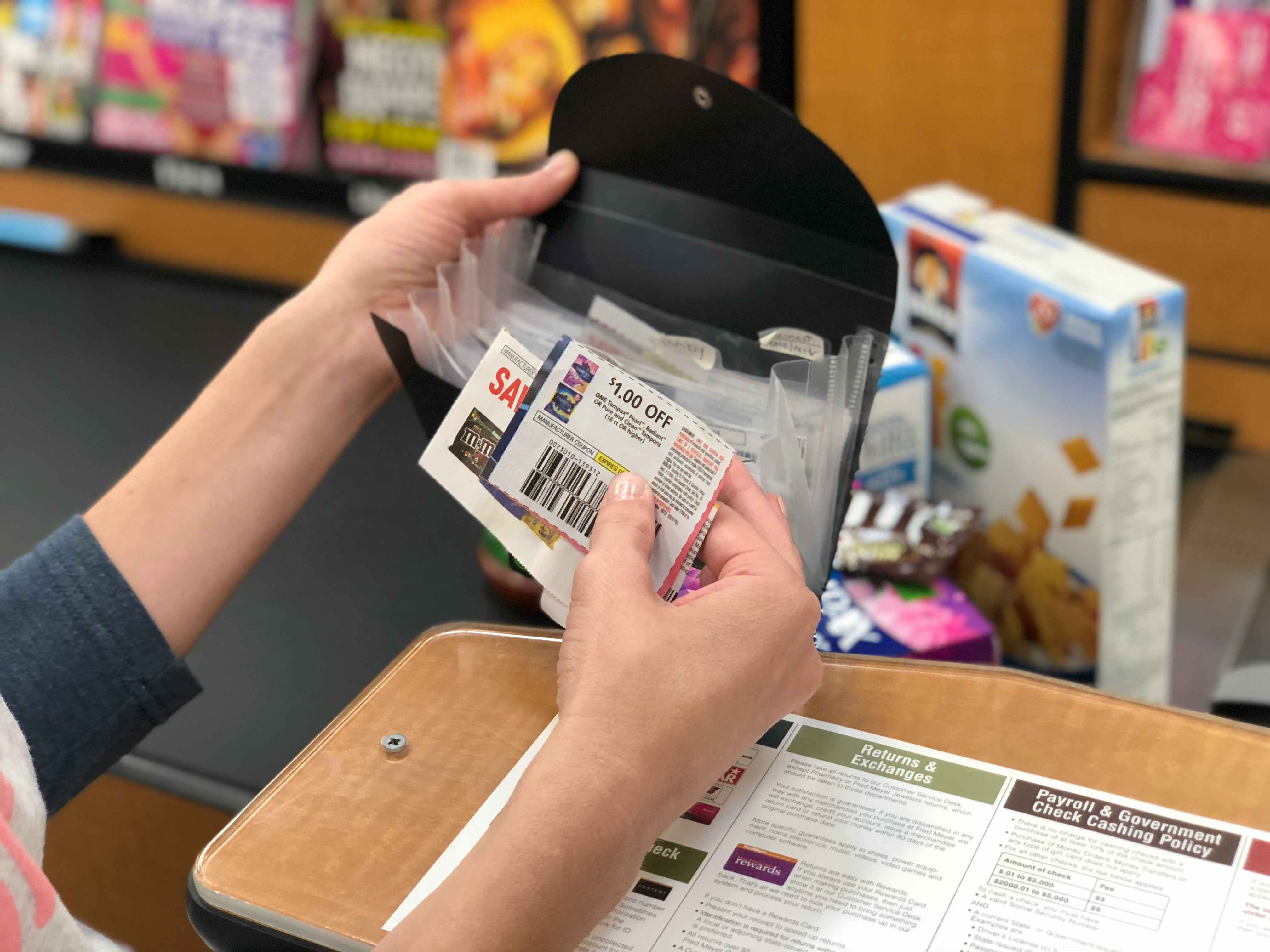 A person's hands holding up a small file organizer and a couple of coupons in front of the conveyer belt of a grocery checkout lane.