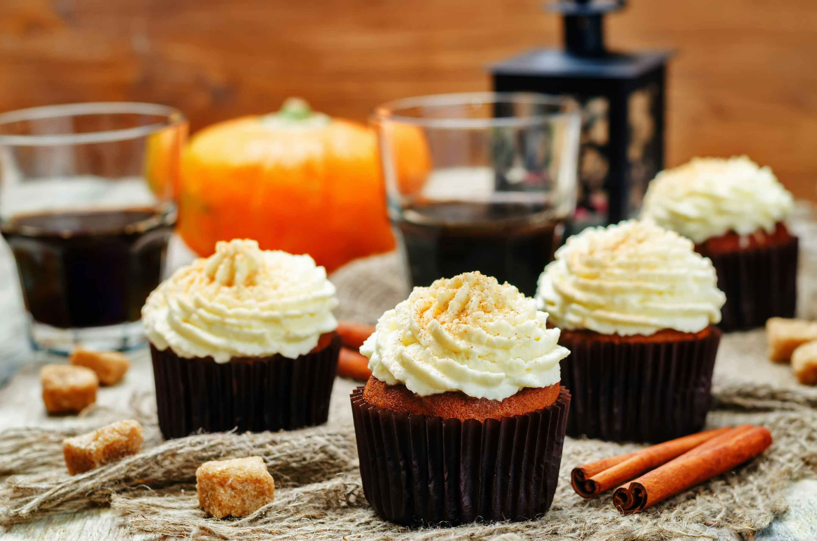 Pumpkin pie cupcakes on a table with drinks, cinnamon sticks, a pumpkin, and some crumbles.