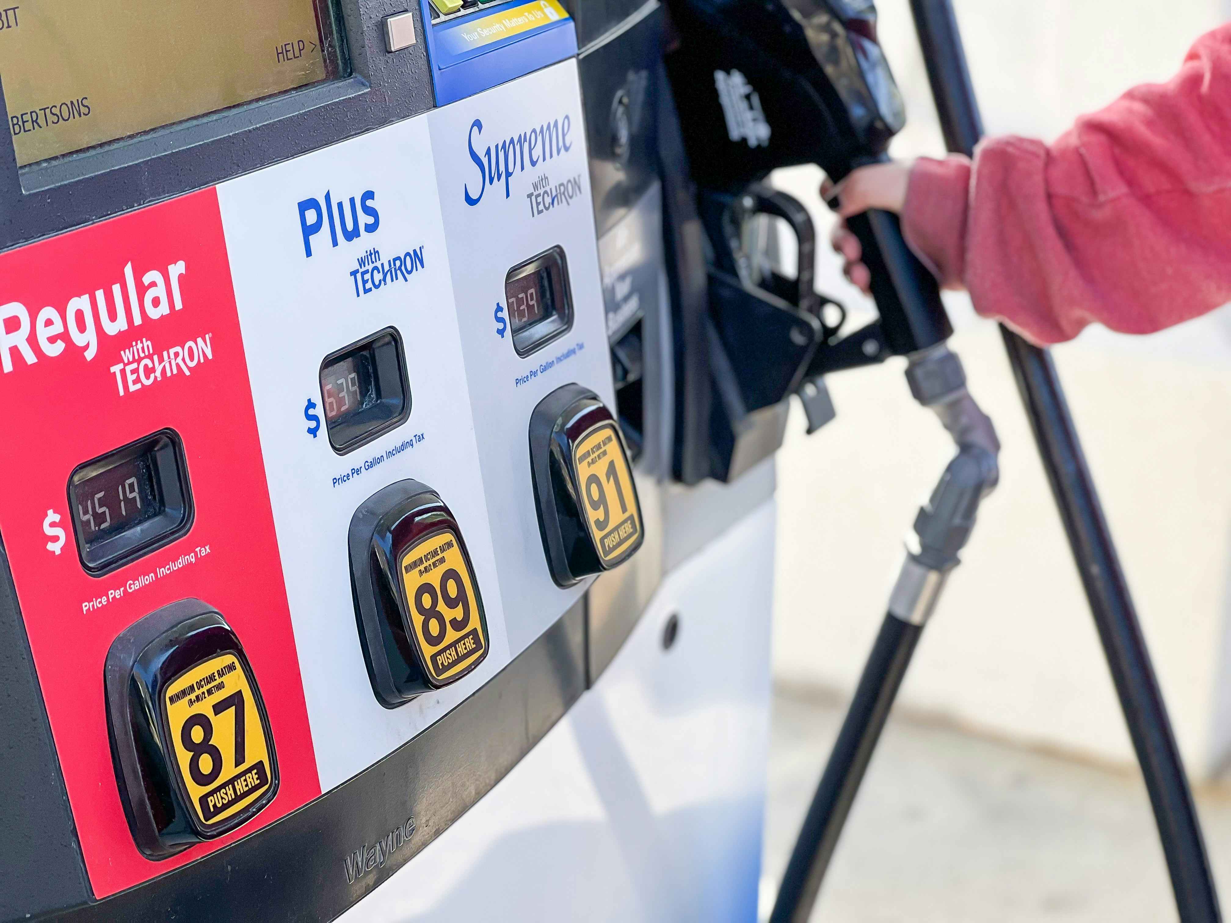 A person's hand reaching to take the nozzle from a gas pump with the fuel option buttons in the foreground.