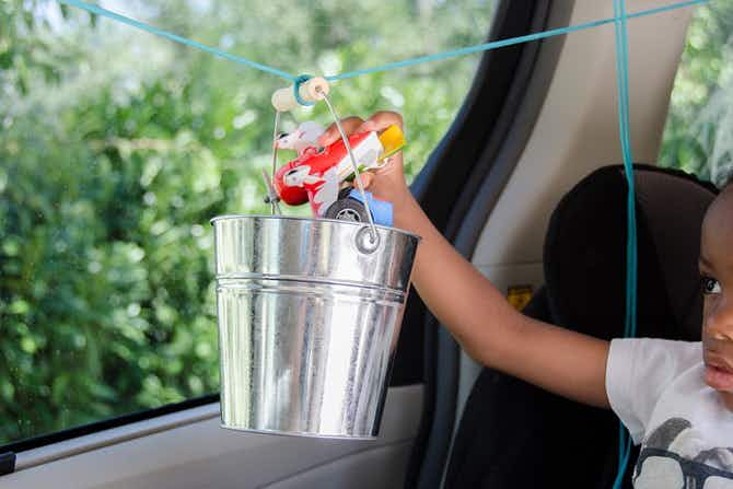 A child sitting in a car, placing a toy airplane into a bucket that is hanging from a string in front of them.