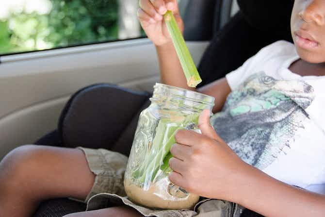 A child sitting in a car, taking a celery stick out of a mason jar filled with more celery sticks and some peanut butter.