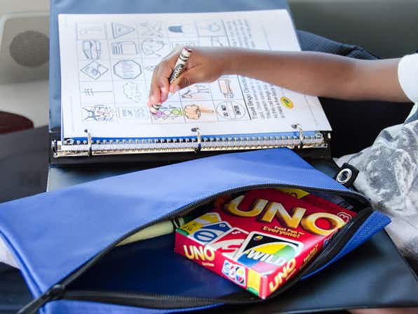 A child using a crayon to color on a printed activity page that is fastened into a binder. A pencil case with more crayons and a pack of Uno playing cards is sitting next to the child.