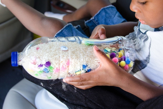A child sitting in a car, looking into a clear plastic water bottle that has been filled with rice and various trinkets to play iSpy.