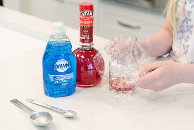 Someone placing plastic wrap over a wine carafe with red wine vinegar and Dawn soap nearby