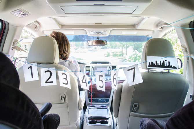 A string of papers with numbers 1-7 printed on them, and one a silhouette of a city, hanging between the driver's and passenger's seat in a van, facing the backseat. A person is sitting in the driver's seat looking forward out of the windshield. 