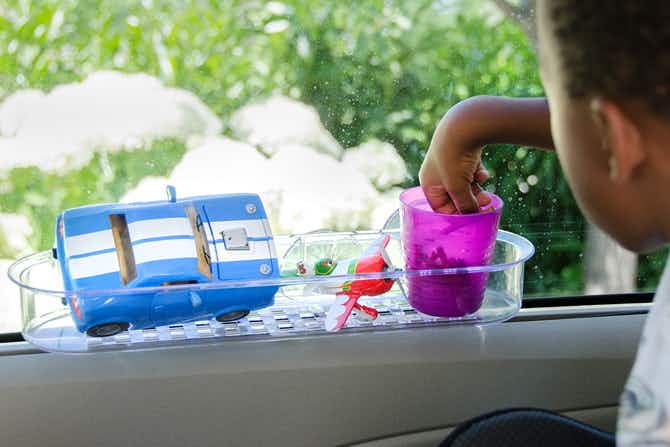 A child reaching into a plastic cup that is sitting in a shower caddy shelf that has been suctioned to the inside window of the car. There is also a toy car and a toy airplane in the shower caddy next to the cup.