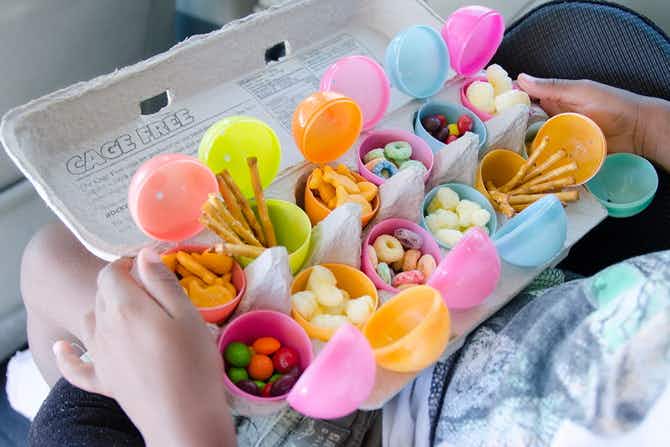 A child sitting in a car holding a cardboard egg carton filled with opened plastic Easter eggs on their lap. Each egg is filled with a different type of snack or candy.