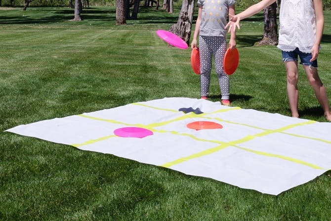 Two kids playing tic-tac-toe with frisbees on a shower curtain liner decorated with tape in a backyard.