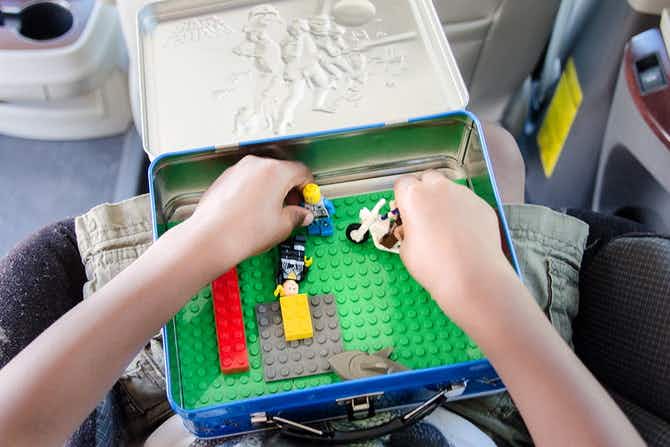 A child's hands playing with LEGO toys inside of a metal lunchbox sitting on the child's lap in the car.