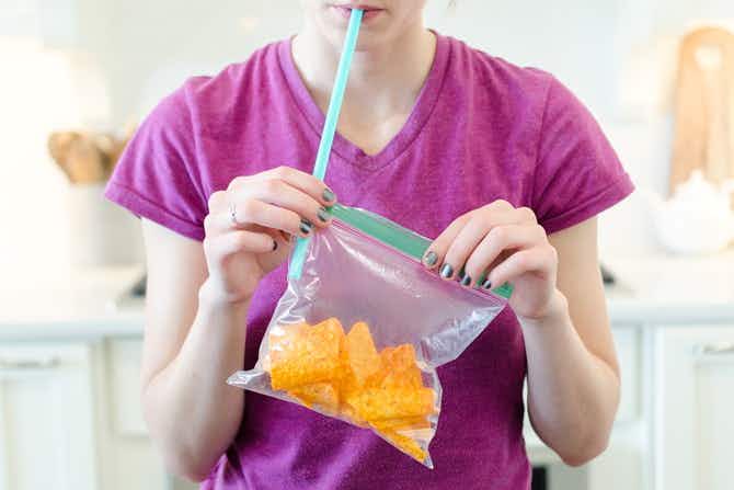 A woman using a straw to blow air into a ziploc bag filled with chips.