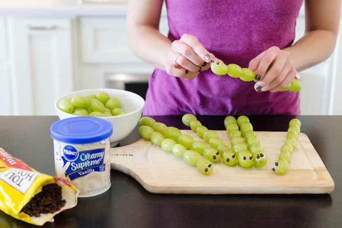 A person making grape caterpillars using grapes, skewers, icing, and chocolate chips.