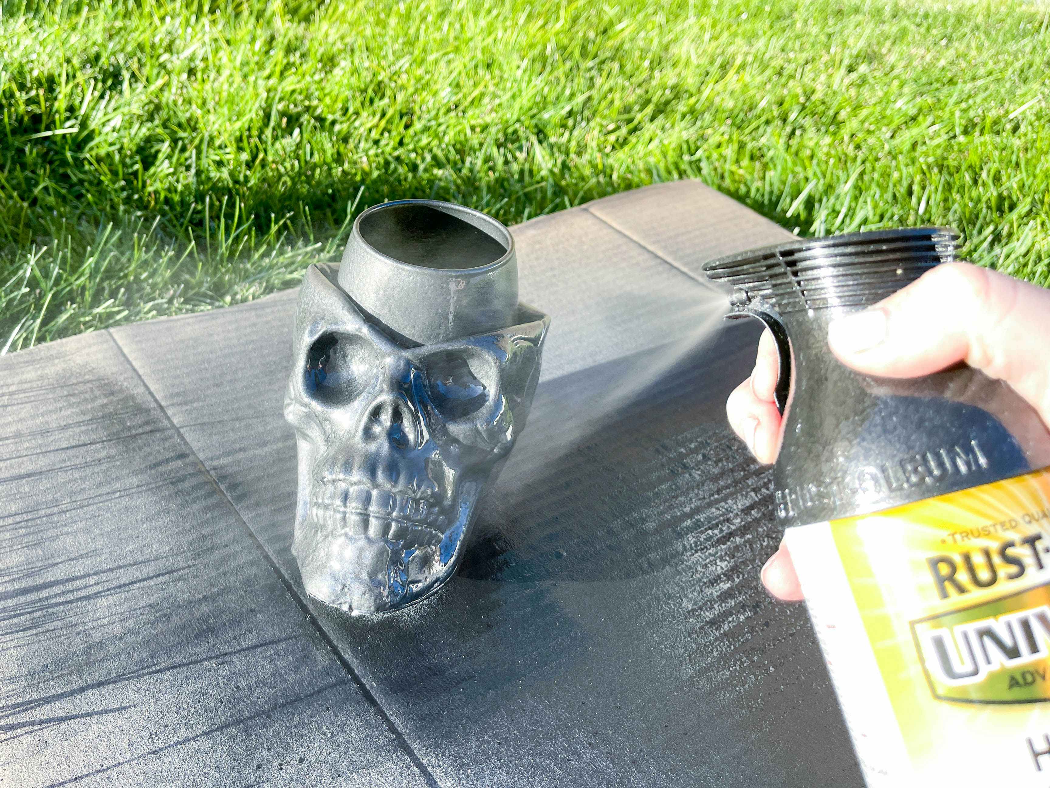 A plastic skull with a glass in the top being spray painted outside on a cardboard piece