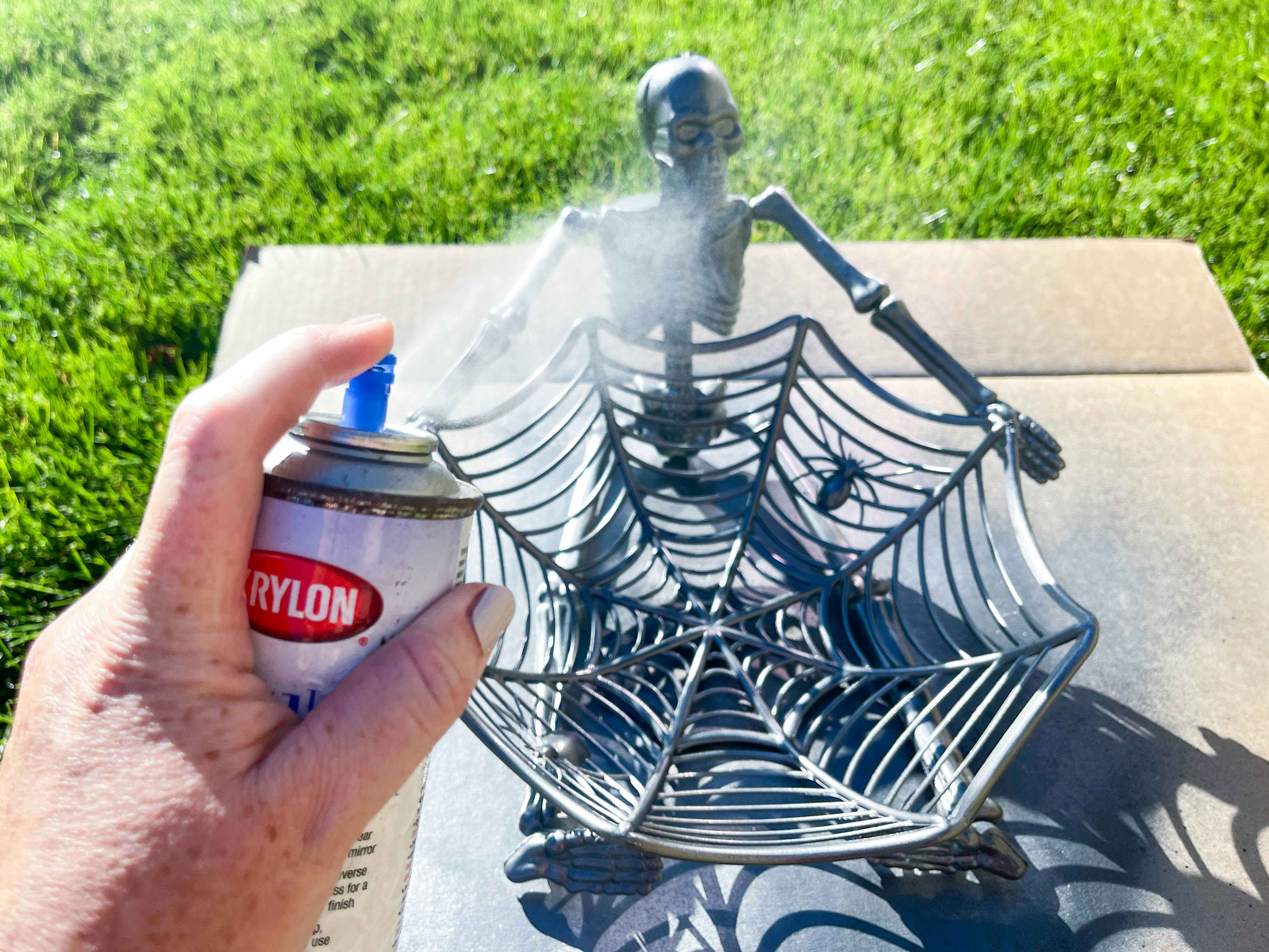 A plastic skeleton glued to a spider web candy bowl being spray painted outside on a piece of cardboard