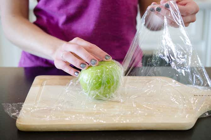 A sliced apple on a cutting board being wrapped in plastic wrap.