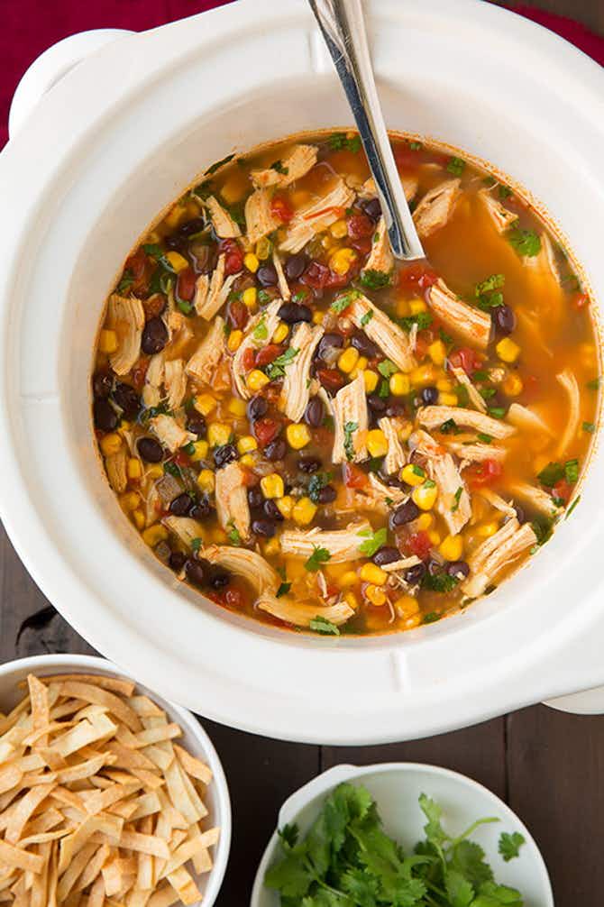 A crockpot soup with chicken, beans, corn and toppings nearby 