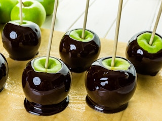 Apples dipped in black toffee candy with bamboo skewers sticking out of them, sitting on a counter.