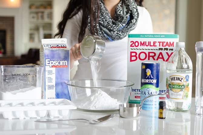 a person pouring borax into a mixing bowl surrounded by homemade dishwasher tabs ingredients 