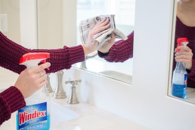 A person holding a bottle of Windex and wiping a mirror with a newspaper.