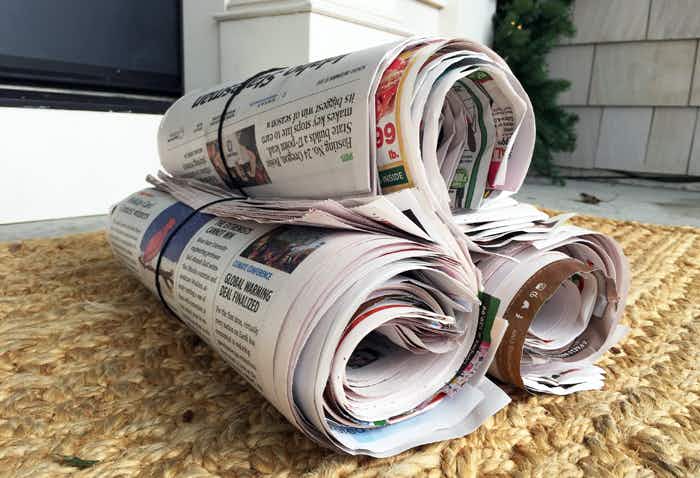 Three newspapers rolled up sitting on top of each other on a rug on a porch in front of house.