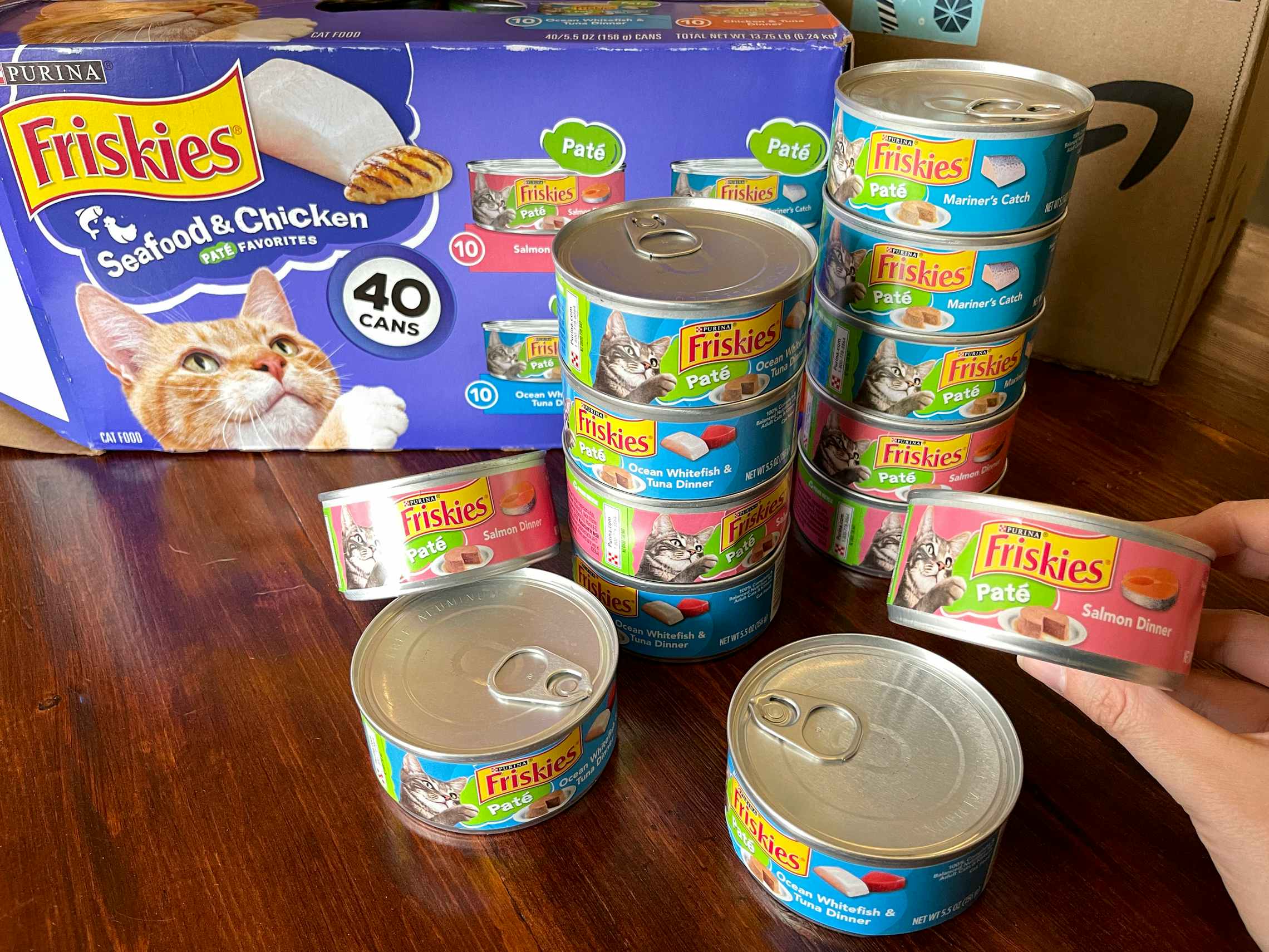 A person's hand holding up a can of Friskies wet cat food next to a stack of more Friskies cans and their box.