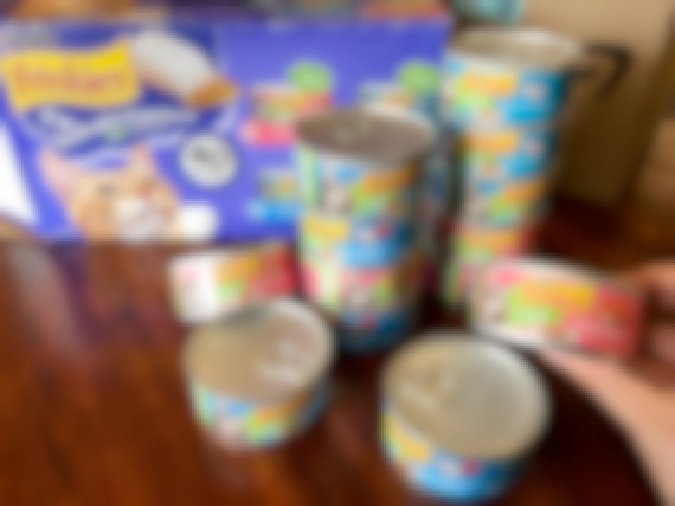 A person's hand holding up a can of Friskies wet cat food next to a stack of more Friskies cans and their box.