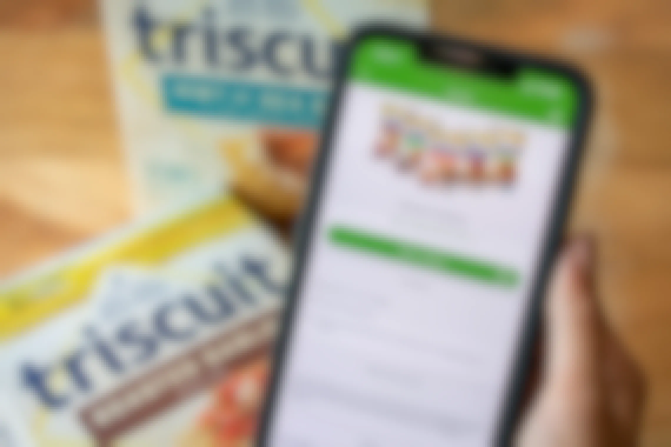 A person's hand holding an iPhone displaying the Checkout 51 mobile rebate app and an offer for Triscuit crackers that the user has claimed. In the background there are two boxes of Triscuit crackers sitting on the table.
