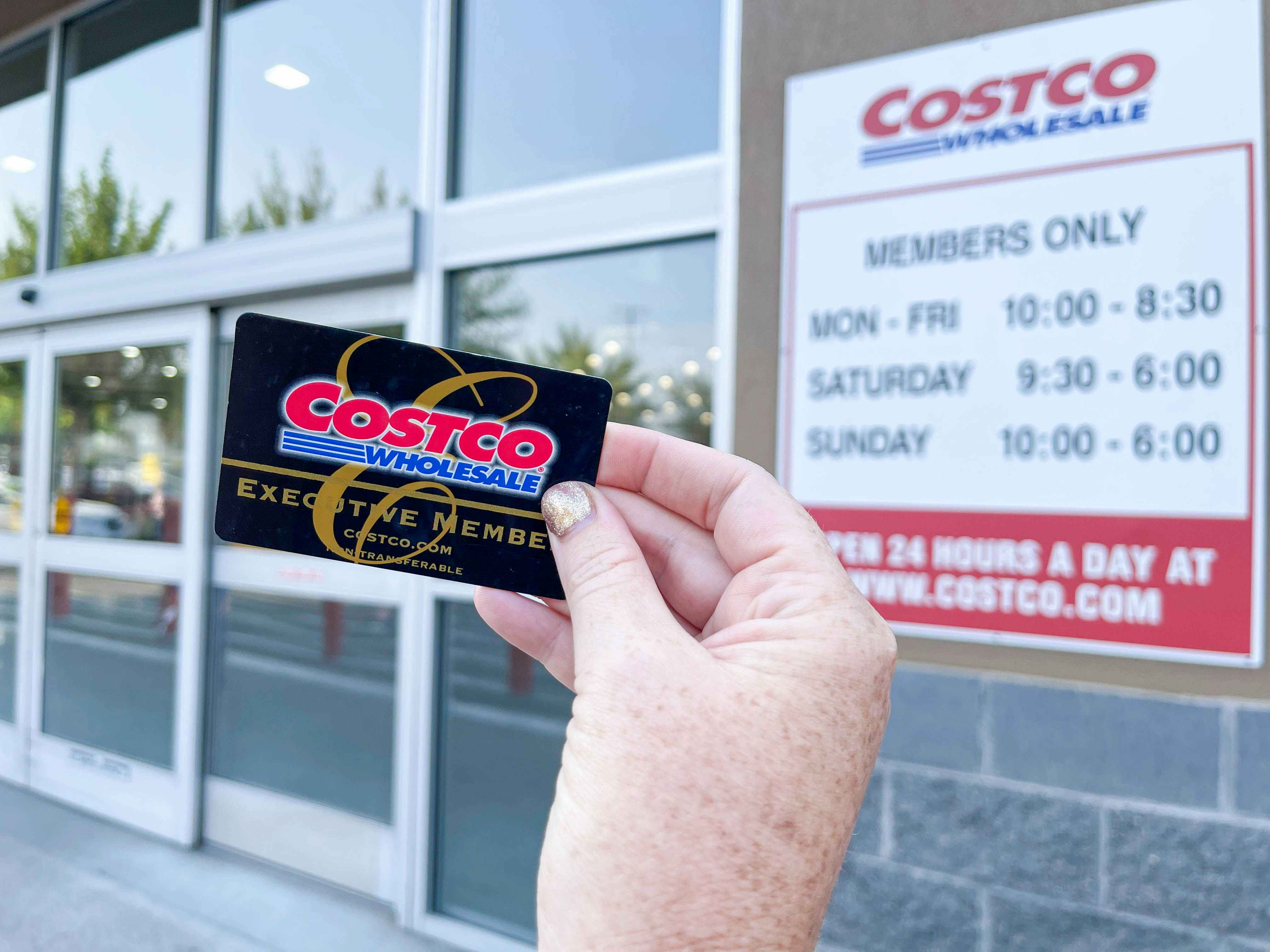 A Costco Membership card being held in front of the Costco store hours sign on the outside of the building.