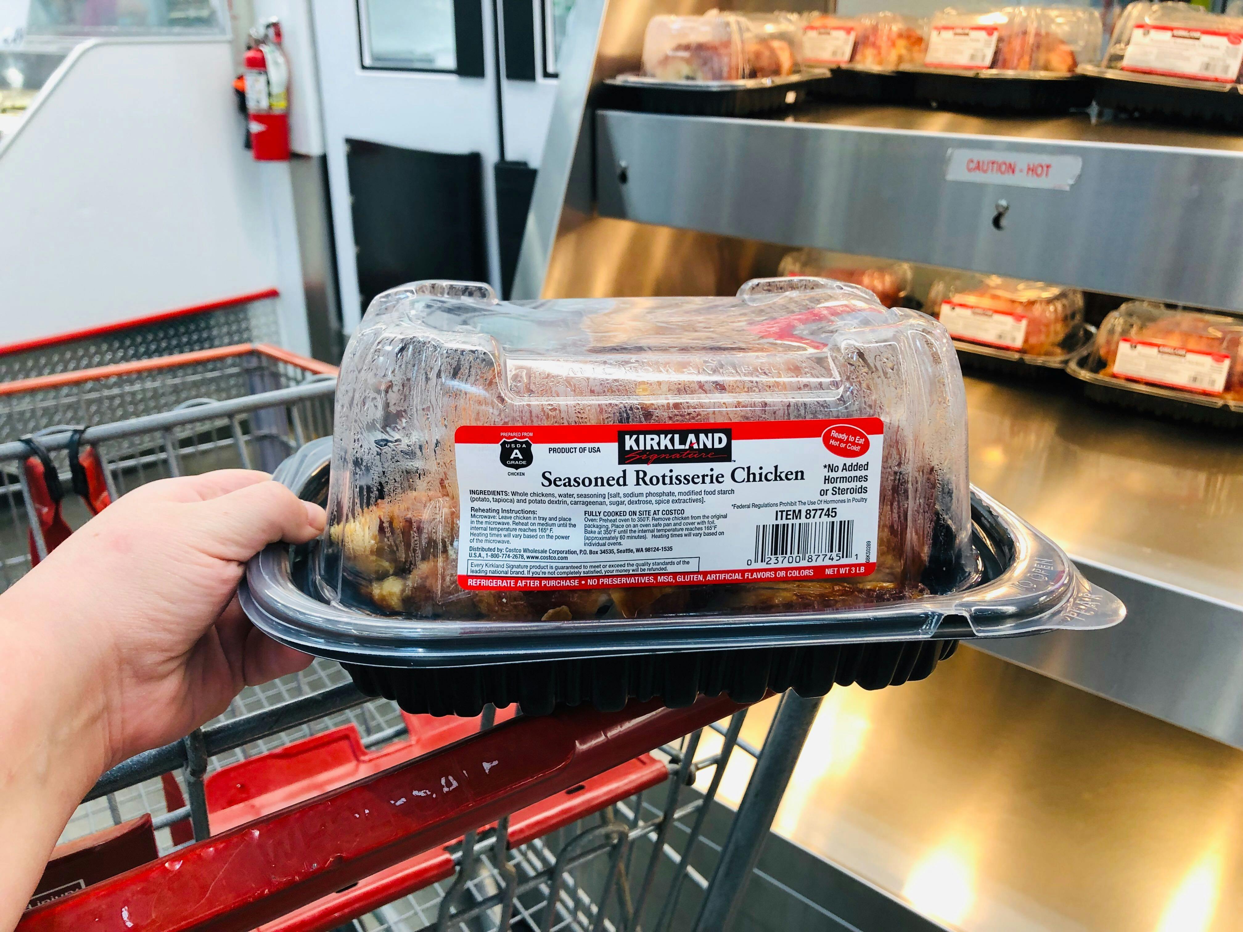 Person holding Costco rotisserie chicken on the edge of a Costco shopping cart.