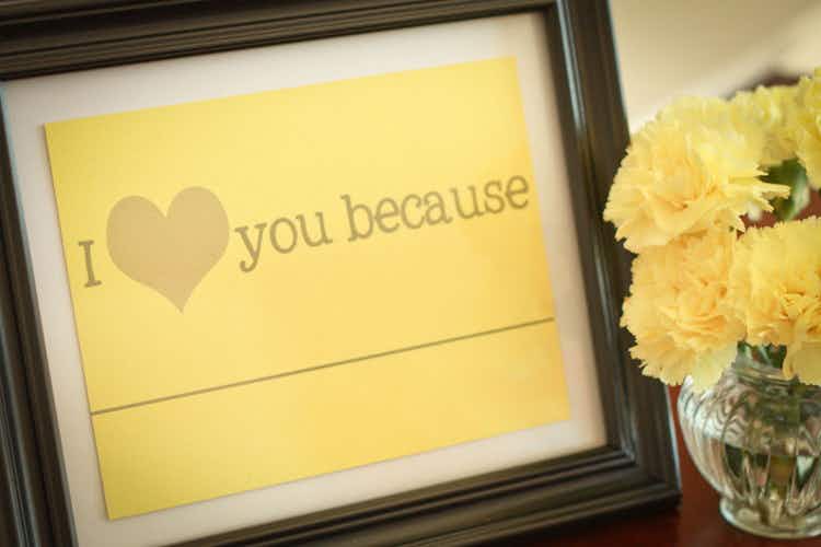 A framed paper that says "I heart you because..." next to a vase of flowers