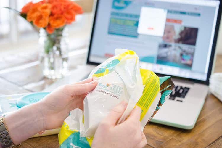 A person looking at the code on the inside of a pampers diapers package. A laptop computer is in the background with pampers website open on the screen