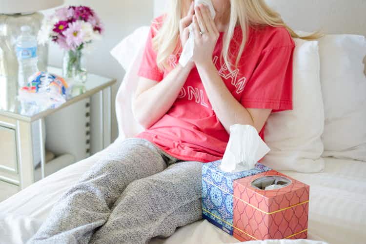 person in bed blowing nose with two tissue boxes nearby