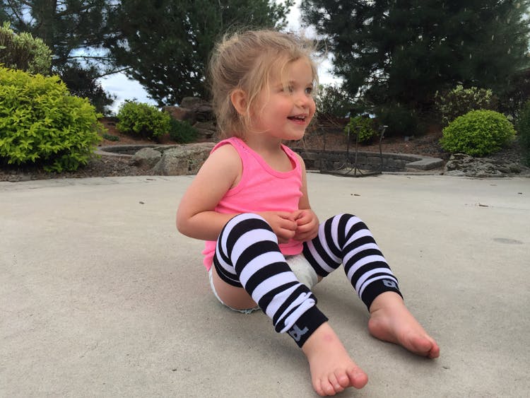 A toddler girl wearing white and black baby leggings while sitting on some pavement outside.