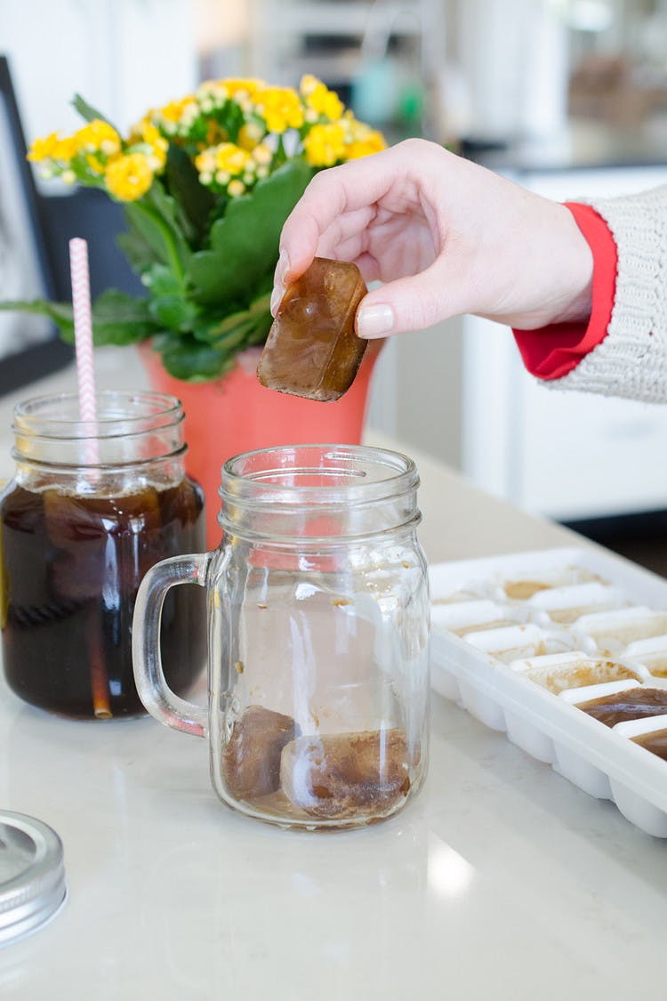 A person putting a coffee ice cube into a glass drinking cup next to an iced coffee with a straw and a tray of more coffee cubes.