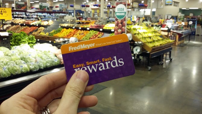 A person holding a Fredmeyer reward card while standing in the produce section of a grocery store.