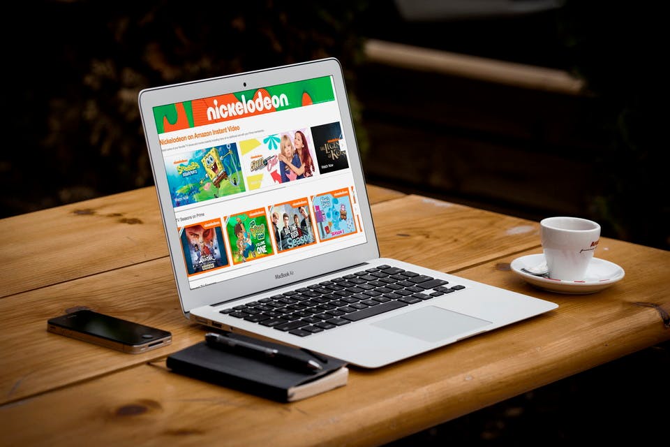 A laptop open on a table displaying a web page that features Nickelodeon shows. Sitting next to the laptop is a notebook and pen, a cellphone, and a small mug.