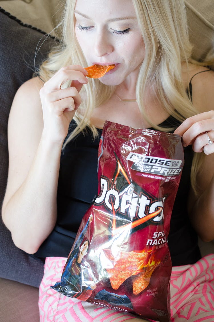 A woman sitting on a couch, holding a bag of Doritos Spicy Nacho chips and putting a chip into her mouth.