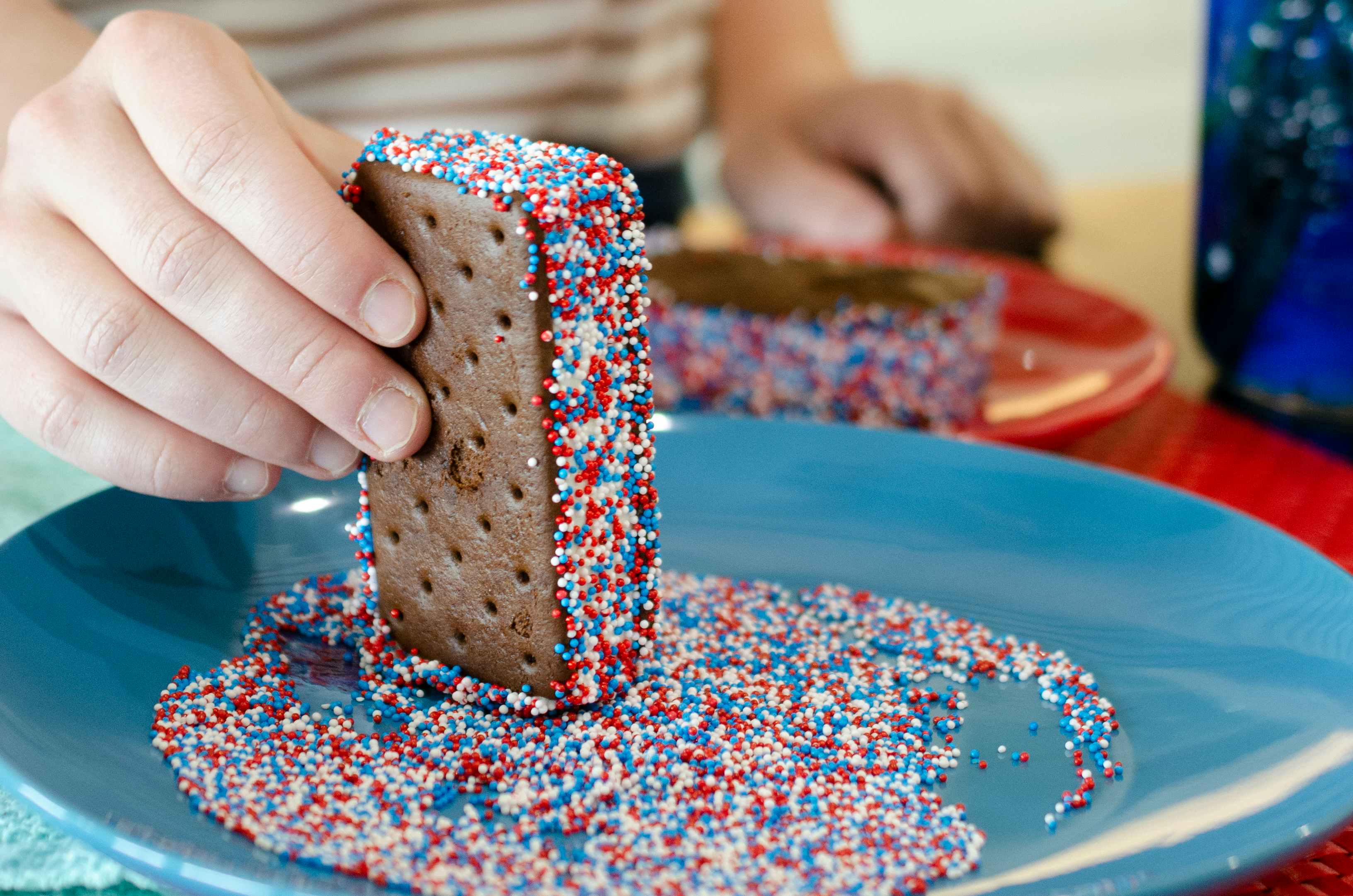 A person dipping an ice cream sandwich in red, white, and blue sprinkles.