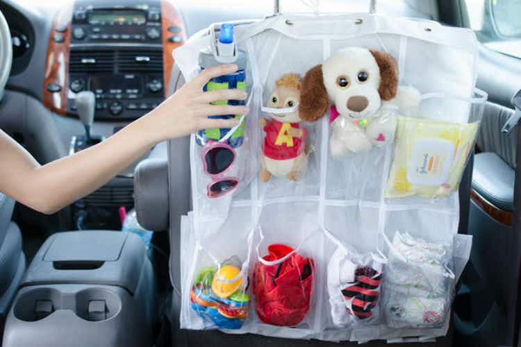 A person's arm reaching from the driver's seat in a car, to put a child's water bottle into a compartment of a shoe organizer that is hanging on the back of the passenger seat. The other compartments are filled with toys, wipes, and diapers.