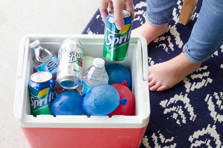 A person's hand picking up a can of soda from a cooler filled with drinks and frozen water balloons.