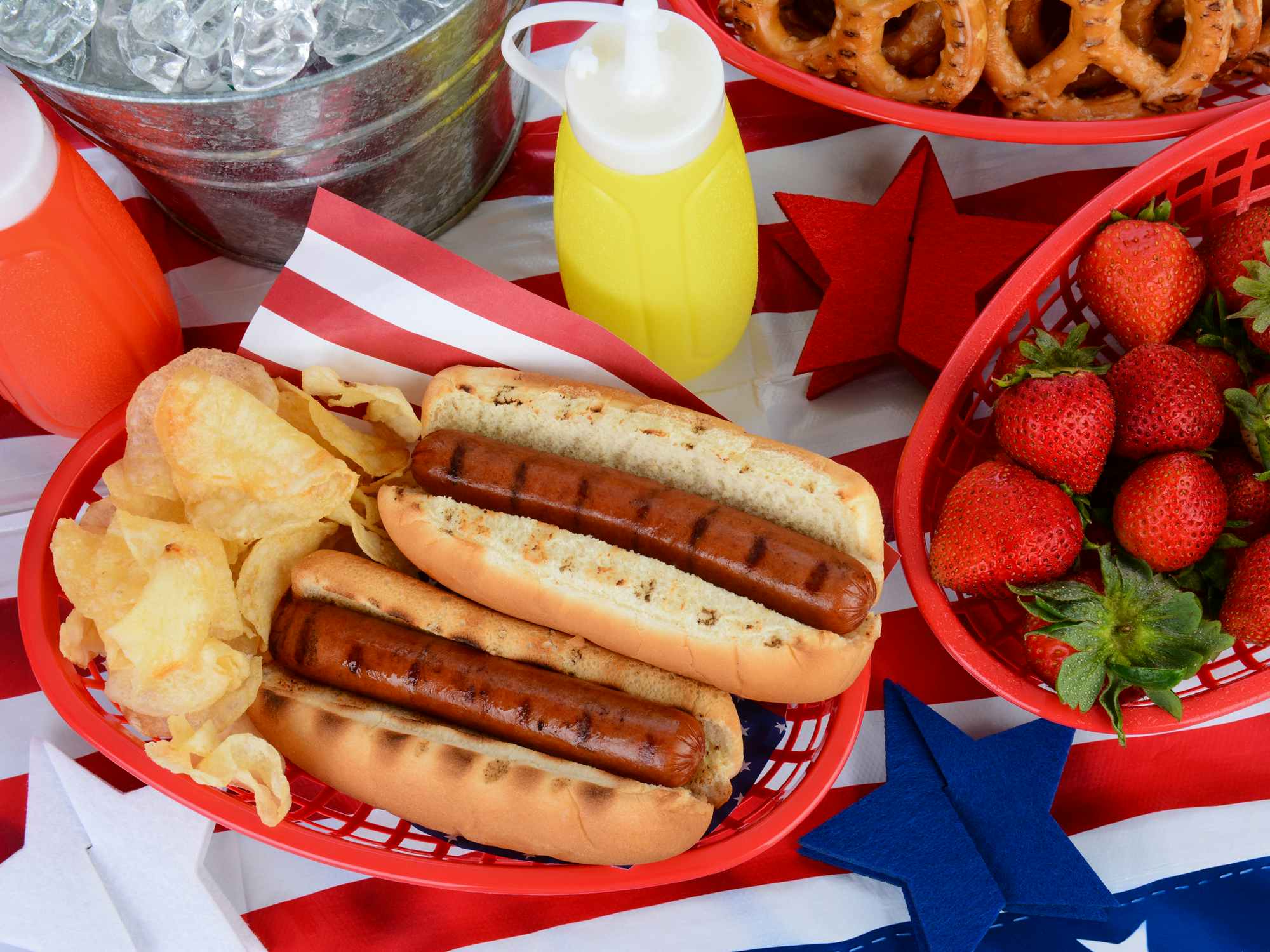 Hot Dogs, chips, strawberries, mustard, ketchup, pretzels, and ice bucket on a table decorated for the 4th of July