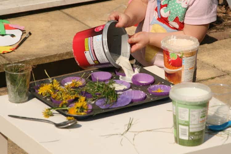a child pouring goop into baking cups on a cooking sheet outside with flowers and grass