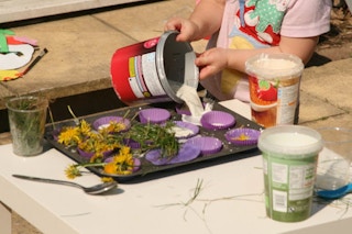 a child pouring goop into baking cups on a cooking sheet outside with flowers and grass